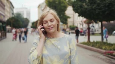 Sokakta yürürken konuşan orta yaşlı sarışın kadının portresi. Modern akıllı telefon teknolojisi. İletişim meşgul.