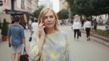 Çekici orta yaşlı sarışın kadın telefonda konuşuyor. Şehir merkezinde yürürken modern akıllı telefon teknolojisi çalışıyor. İletişim özgüveniyle ilgili portreyi kapat.