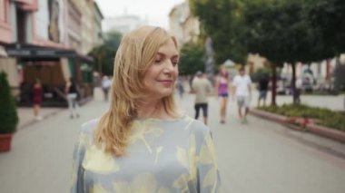 Orta yaşlı sarışın kadın kameraya bakıyor. Şehir merkezinde gülümsüyor. Son sınıf, çekici, dış görünüşü güzel.