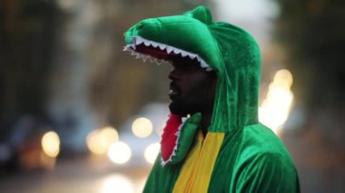Komik dinazor genç Afro-Amerikalı gülümseyen yeşil T-rex oynayan adam yağmurlu bir akşamda şehir merkezinde caddede duruyor..
