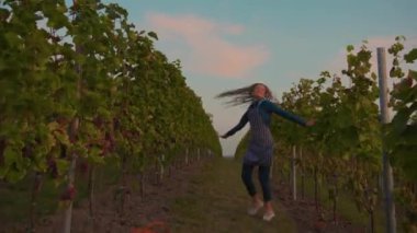 Şık genç çiftçi kadın üzüm bağında zıplayarak dans ediyor. Güneşli bir sonbahar günü gülümsüyor. Mutlu tarım endüstrisi, asma tarlası, taze meyve, yeşil eller yavaş çekim işçisi.