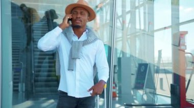 Şapka standında bavullu yakışıklı Afro-Amerikan havaalanı yakınlarında telefonla konuşuyor. Tatil ve seyahat. Sohbet başarılı bir açık hava stili gezisi. Kapatın. Yavaş çekim