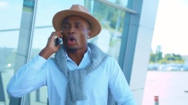 Şapka standındaki sinirli, yakışıklı Afro-Amerikan havaalanı yakınlarında telefonda konuşuyor. Tatil ve seyahat. Açık havada güzel bir sohbet. Kapatın. Yavaş çekim