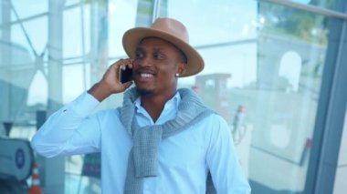 Şapka standında bavul taşıyan yakışıklı Afro-Amerikalı havaalanı yakınlarında telefonda gülümsüyor. Tatil ve seyahat. Sohbet başarılı bir açık hava stili gezisi. Kapatın. Yavaş çekim