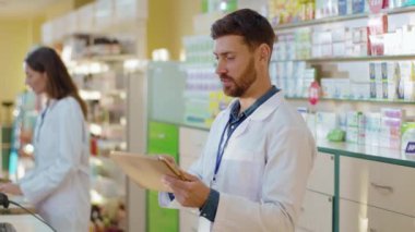 Kafkasyalı erkek eczane stajyeri dijital tabletle ilaçları araştırıyor eczanede çalışan profesyonel eczacıdan tavsiye istiyor. Sağlık ve İlaç Hizmetleri.