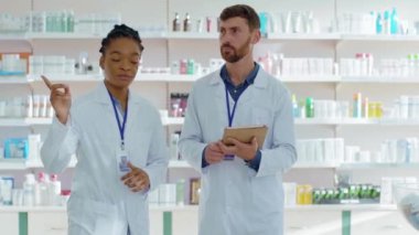 Afrika kökenli Amerikalı kadın eczacı, eczane sağlık merkezindeki ilaç ve ilaçları inceleyen bir tablet bilgisayar kullanarak erkek stajyere danışmanlık yapıyor. İşbirliği. Karışık ırk.