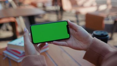 NEW YORK - 5 Nisan 2019: Kafede yatay yeşil ekranlı akıllı telefon kullanan, el ele tutuşan genç kadın açık havada yeni teknolojiyi yavaş çekimde görüntülüyor.