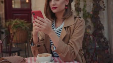 Yüzünü kapat. Kırmızı ruj şapkalı, şık bir ceket giyen, güneşli bir günde telefonda yazan, kahvaltının üzerinde oturan, açık havada gülümseyen, şehir modasına uygun, yavaş çekim yapan genç bir kadın.