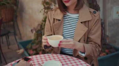 Kahvaltının üzerinde bir fincan kahveyle masada oturan genç bir kadın. Sabah kruvasan modayla kameraya bak. Gülümse.