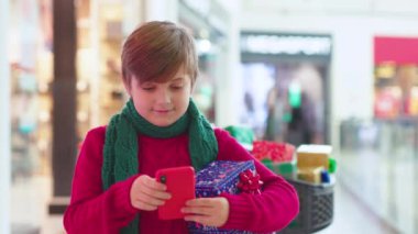Çocuk elinde Noel hediyesi kutularıyla alışveriş merkezinde duruyor. Telefon, aile, çekici ergen çocuklar, neşeli, mutlu, mutlu bir tatil çocuğu, çocukluğunda ağır çekimde gülümsüyor.