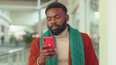 Mutlu Afrikalı Amerikalı genç adam, Noel zamanı AVM 'de telefon yürüyüşü yapıyor. Noel ağacı gülümsüyor, gülümsüyor, internete bakıyor, mobil portre teknolojisini kapatıyor.