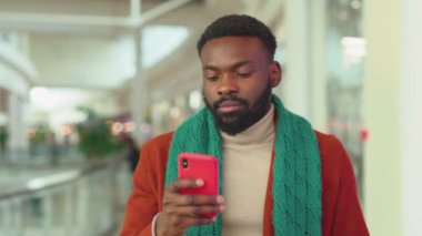 Ciddi Afrikalı Amerikalı genç adam Noel zamanı AVM 'de telefon yürüyüşü yapıyor. Noel ağacı gülümsüyor, gülümsüyor, internete bakıyor. Yavaş çekim mobil portre teknolojisini kapatın.