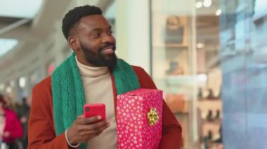 Noel hediyesi olan Afrikalı Amerikalı genç adam alışveriş merkezinin arka planında telefon yürüyüşü yapıyor. Noel ağacı gülümsüyor, internete gülümsüyor. Mobil teknoloji hücresinin portresini yavaş çekimde kapatın.