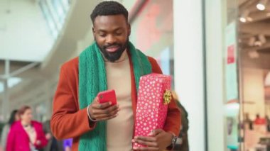 Ağır çekimde Afrikalı Amerikalı genç adam Noel zamanı AVM 'de telefon yürüyüşü yapıyor. Noel ağacı gülümsüyor, internete bakıyor, portre teknolojisini yakından görüyor.