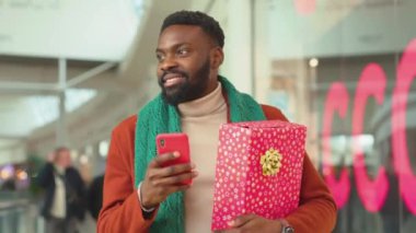 NEW YORK - 5 Nisan 2019: Yakışıklı Afro-Amerikan genç adam Noel zamanı AVM arka planında telefon yürüyüşü yapıyor Noel ağacı mutlu alışveriş interneti yüzleri mobil portre teknolojisi