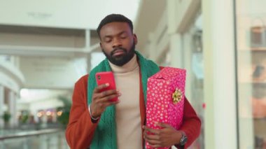 Ciddi Afrikalı Amerikalı adam Noel hediyesi olarak alışveriş merkezinde telefon yürüyüşü yapıyor. Noel ağacı gülümsüyor, internete gülümsüyor ve portre portresini yavaş çekimde çekiyor.