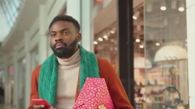 Ağır çekimde Afrikalı Amerikalı genç adam telefon yürüyüşü yapıyor Noel zamanı arka planda Noel ağacı mutlu alışveriş yüzleri yakın portre mobil teknolojisi