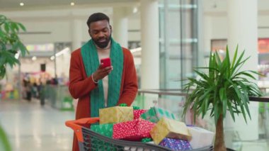 Noel hediye kutularını el arabasında taşıyan Afrikalı Amerikalı genç adam alışveriş merkezinin arka planında telefon kulübesini kullanırken mutlu ol. Gülümse, gülümse, gülümse, gülümse.