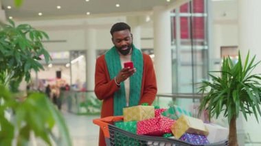 Mutlu Afro-Amerikan genç adam Noel hediyesi kutularıyla alışveriş merkezinde telefon kulübesi kullanıyor gülümsüyor, internetin yüzünü kapıyor mobil teknoloji hücresi yavaş çekim portresini çekiyor.