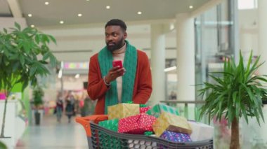 Portre Afro-Amerikan genç adam Noel hediye kutularıyla alışveriş merkezinde telefon kulübesi kullanıyor gülümsüyor, internetin yüzüne gülümsüyor mobil portre teknolojisini yavaş çekimde kapatıyor.