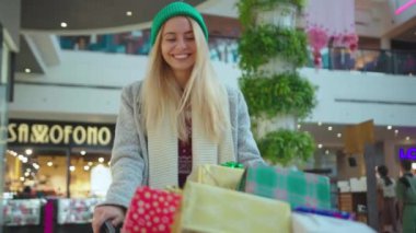 Çekici, genç, çekici, sarışın bir kadın Noel hediyeleriyle alışveriş arabasında kutular içinde geziyor, gülümsüyor, alışveriş yapıyor, yeni yılı yakından izliyor, yavaş çekimde yakın çekimde tatil yapıyor.