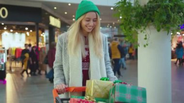 Şık, genç ve çekici bir kadın Noel hediyeleriyle yürüyor alışveriş merkezinde kutular içinde alışveriş yaparken gülümsüyor, alışverişe çıkıyor ve yeni yılı yakından alıyor yavaş çekimde yakın çekimde tatil yapıyor.