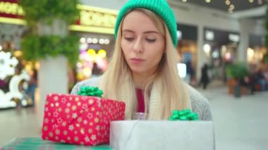 Genç ve güzel bir kadın Noel hediyeleriyle yürürken şapka takıyor. Alışveriş merkezinde kutuları yere düşürmeye çalışıyor. Üzücü yeni yıl tatili portresi alın.