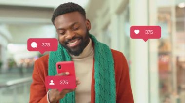 Alışveriş merkezinde Noel hediye kutularıyla gülümseyen çekici Afro-Amerikalı genç adam akıllı telefon kullanarak yorumcu sayacı blogcu yavaş çekim blogcusu gibi sosyal medya simgeleri yazıyor.