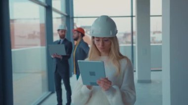 Ciddi kadın emlakçı kask takıyor. Kameraya tablet bilgisayarla bakıyor. Yeni apartman boş.