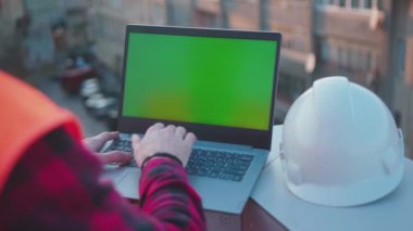Güzel bir arka plan mühendisi balkon kask işçisi projesinde yeşil ekranlı dizüstü bilgisayar kullanıyor. Profesyonel erkek güvenlik bilgisayarı yavaş çekimde modelleme yapıyor.