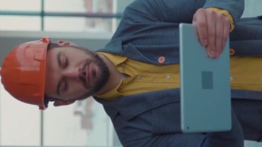 Dikey portre videolu emlakçı kask takıyor. Kameraya tablet bilgisayarla bakıyor. Yeni apartman dairesi boş.