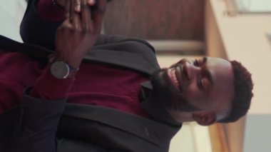 Portre gülümseyen Afrikalı Amerikalı genç adam güneşli bir günde akıllı telefon standı kullanıyor. Yakışıklı yolcular internetteki dikey video ekranı yavaş çekim tarzında daktilo çekiyor.