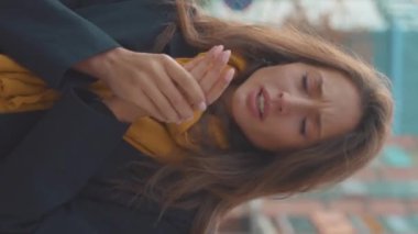 Dikey videoda genç bir kadın hapşırıyor. Açık havada ateşi var. Soğuk alerji şehri. Güzel, hastalıklı, burnu akan kadın.