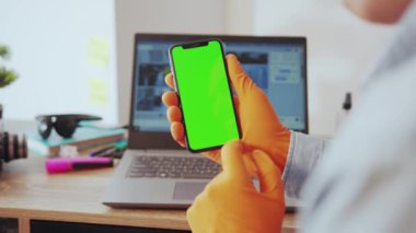 NEW YORK - 28 Mart 2020: Genç bir iş adamı evinde bilgisayarla çalışan dikey krom anahtar modelleme ekranında akıllı telefon kullanıyor. Evdeki insanlar. Teknoloji. Karantina.