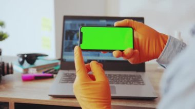 NEW YORK - 28 Mart 2020: Akıllı telefon yeşil ekran uygulaması ile okuyan genç adam yatay poziyonda duruyor evde bilgisayarın başında oturuyor. İnsanlar, teknoloji. Sağlık hizmetleri.