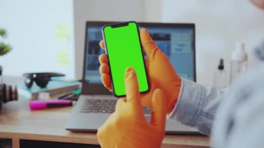 NEW YORK - 28 Mart 2020: Genç bir adam elinde bir akıllı telefonu tutuyor ve yeşil maket ekranda eldivenlerini yazarak evden serbest çalışıyor. Yakın plan. İnsanlar, teknoloji. Karantina.