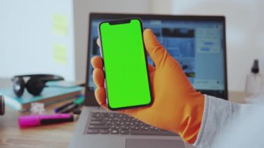 NEW YORK - 28 Mart 2020: Eldivenli bir adamın dikey akıllı telefon dinleme ve evde dizüstü bilgisayarda çalışmak için yeşil ekran mobil uygulama işini kullanması. Karantina, sağlık hizmetleri.