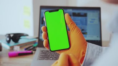 NEW YORK - 28 Mart 2020: Masada oturan akıllı telefon yeşil ekran dikey krom anahtar uygulaması kullanarak evde çalışan bir adamın fotoğrafı. El bilgisayarı, eldiven koruması. Sağlık hizmetleri. Karantina.