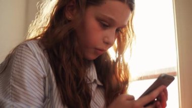 Güzel genç kız pencerenin yanında oturan akıllı telefonu kullanıyor. Mutlu güneş ışığını hisset. Çocuk evinde.