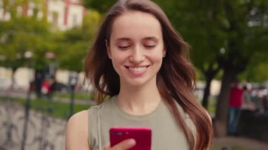 Parlak gün ışığında gülümseyen genç kadın şehir merkezinde yürürken telefon kullanıyor. Yazın internet işi teknolojinin dışında. Yavaş çekim mobil sokak mutlu portresi.