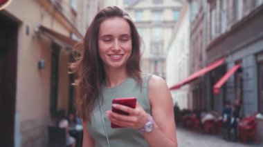 Parlak gün ışığında gülümseyen genç kadın şehir merkezinde yürürken kırmızı telefon kullanıyor. Yazın internet işi teknolojinin dışında.