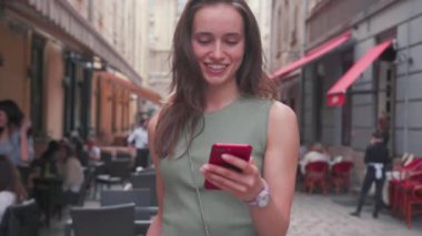 Parlak gün ışığında gülümseyen güzel bir genç kadın şehir merkezinde yürürken kırmızı telefon kullanıyor. Yazın internet işi teknoloji, bahar, mobil, sokak, mutlu portre.