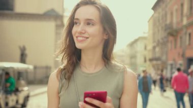 Parlak güneş ışığı altında gülümseyen genç bir kadın şehir merkezinde yürürken kırmızı telefon kullanıyor. Yaz mevsiminde internet sektörüne göz gezdirme teknolojisi dışında gezdirme amaçlı mobil mobil sokak mutlu portresi.