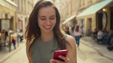 Parlak günışığında gülümseyen genç bir kadın şehir merkezinde yürürken kırmızı telefon kullanıyor. Yazın internet işi teknolojinin dışında.
