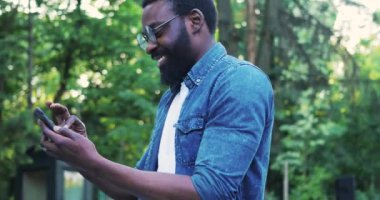 Gözlüklü genç bir Afro-Amerikan adam telefon gülümsemesi kullanır. Sokak güneşinde mutlu hissedin. İnternette teknolojiyle yüzleşin.