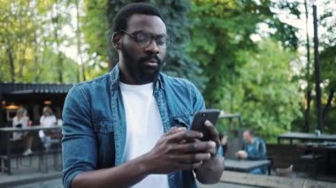 Ciddi Afro-Amerikan gözlüklü genç adam sokak güneşinde telefon kullanıyor. İnternet teknolojisi. Yakışıklı, akıllı telefon mesajları.