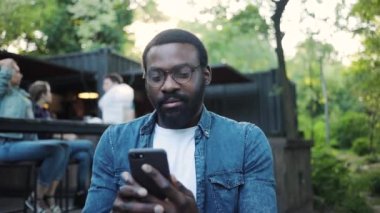 Gülümseyen Afro-Amerikan gözlüklü genç adam telefonu kullan. Telefon kullan. Sokak güneşinde mutlu ol.