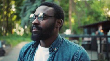 Mutlu yüz, gözlüklü Afro-Amerikalı adam kameraya bakıyor. Gülümse. Teknoloji, doğa, günlük güneş, siyah, yakışıklı, açık hava sakalı, neşeli, kentsel, ağır çekim.