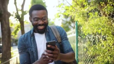 Yavaş çekim mutlu Afro-Amerikalı gözlüklü yakışıklı adam. Sokakta yürür, gülümser, günbatımı gülümser. Cep telefonu moda interneti.