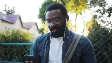 Gözlüklü Afro-Amerikan genç ve çekici bir adam sokakta telefon kulübesi kullanıyor kulaklıkla müzik dinliyor günbatımı gülüşü cep telefonu dansı interneti siyah şehir arabasının dışında.